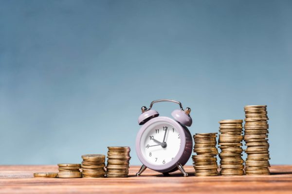 Purple-alarm-clock-between-the-stack-of-increasing-coins-on-wooden-desk-against-blue-background